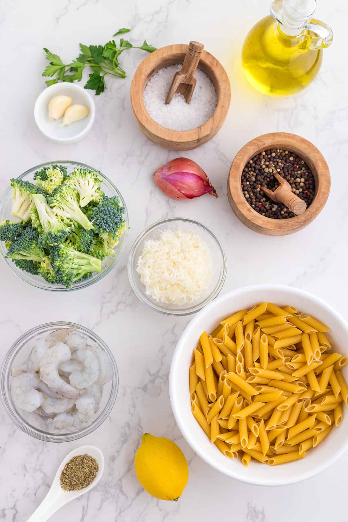 Ingredients for a pasta dish arranged on a white surface, including penne, shrimp, broccoli, garlic, shallot, parmesan, lemon, parsley, olive oil, salt, and pepper.
