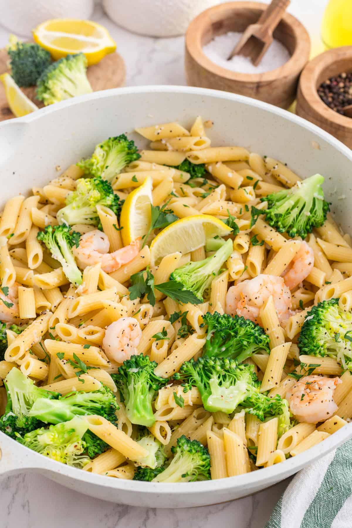 Pasta penne with shrimp, broccoli, lemon slices, and herbs in a white pan, with salt, pepper, and lemon wedges in the background.