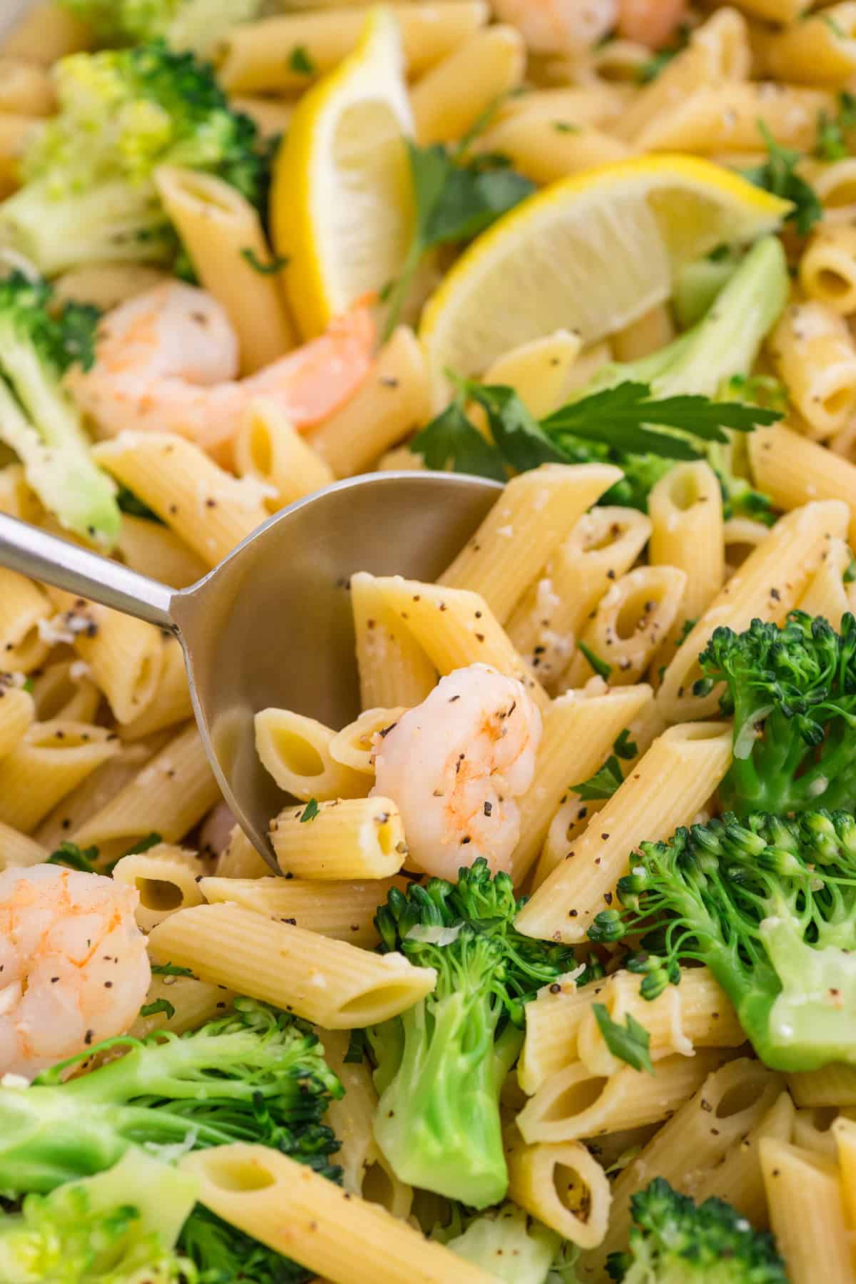 Close-up of penne pasta with shrimp, broccoli, parsley, and lemon wedges, served with a metal spoon.