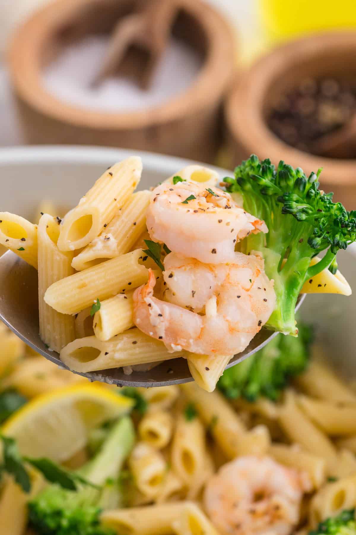 A close-up of a spoon holding penne pasta, shrimp, and broccoli, with a bowl of pasta and wooden containers in the background.