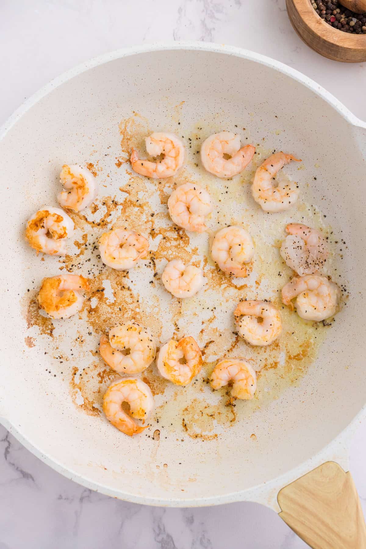 Cooked shrimp with browned bits are scattered in a white skillet on a marble surface, with a wooden bowl of spices visible in the top right corner.