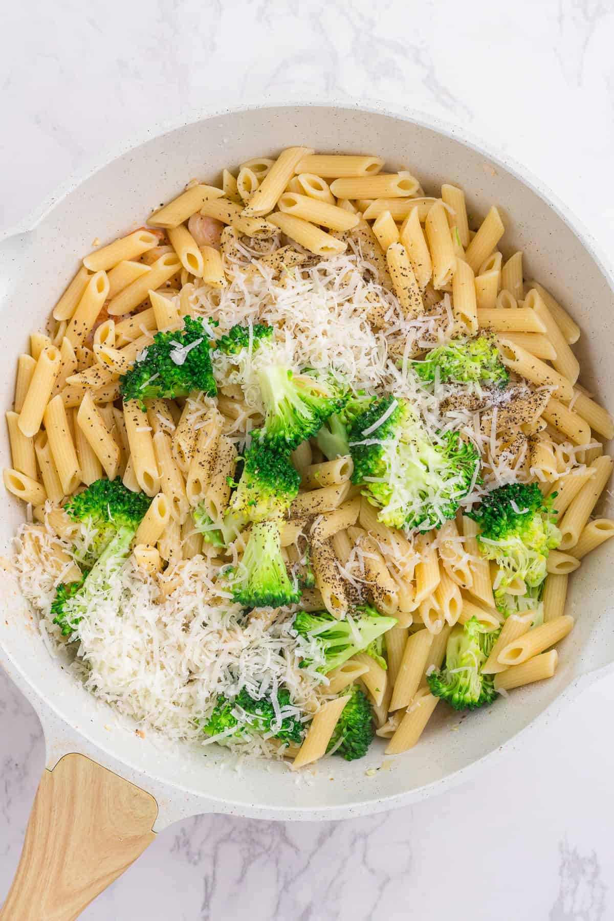 A white skillet with cooked penne pasta, broccoli florets, grated cheese, and black pepper on a marble surface.